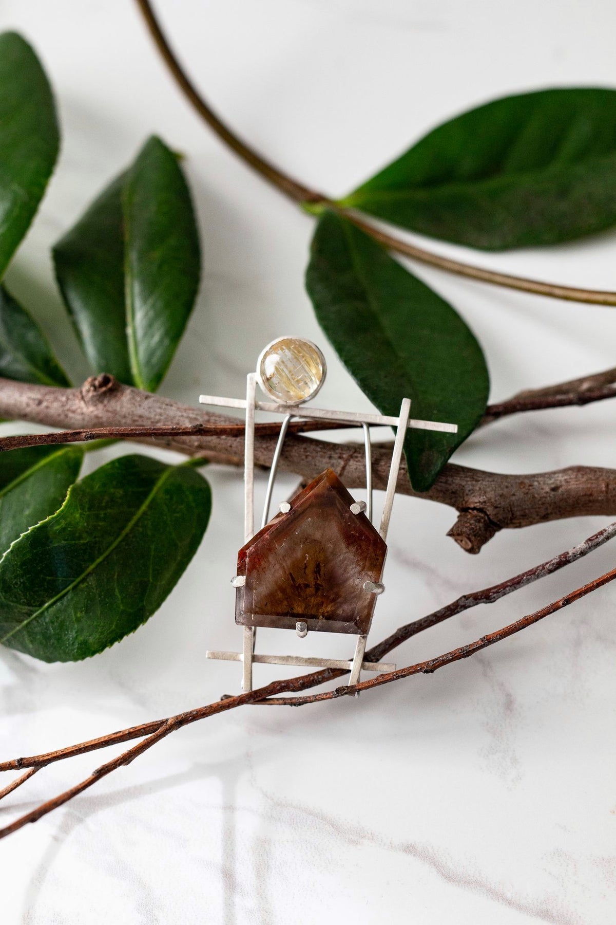 Contemporary architectural sterling silver brooch featuring a rutilated quartz cabochon and a raw super seven quartz gemstone, displayed on natural branches.