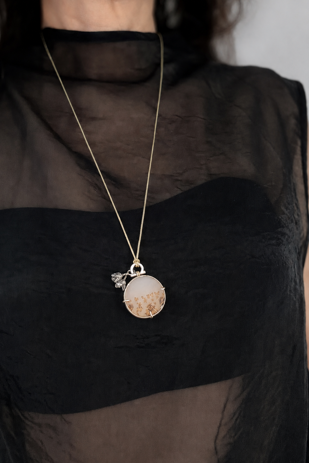 Editorial style shot of the handmade circular dendritic agate pendant necklace worn over a sheer, textured high neck top. The round gemstone showcasing natural amber and brown branch-like inclusions, is suspended long fro, a fine golden silk cord against the layered black fabric. The piece displays its structural silver fixed-arch bail with a 14ct recycled gold wire coil, three delicate prongs, and a dark oxidised cast-from-nature eucalyptus pod charm dangling alongside the silver setting.