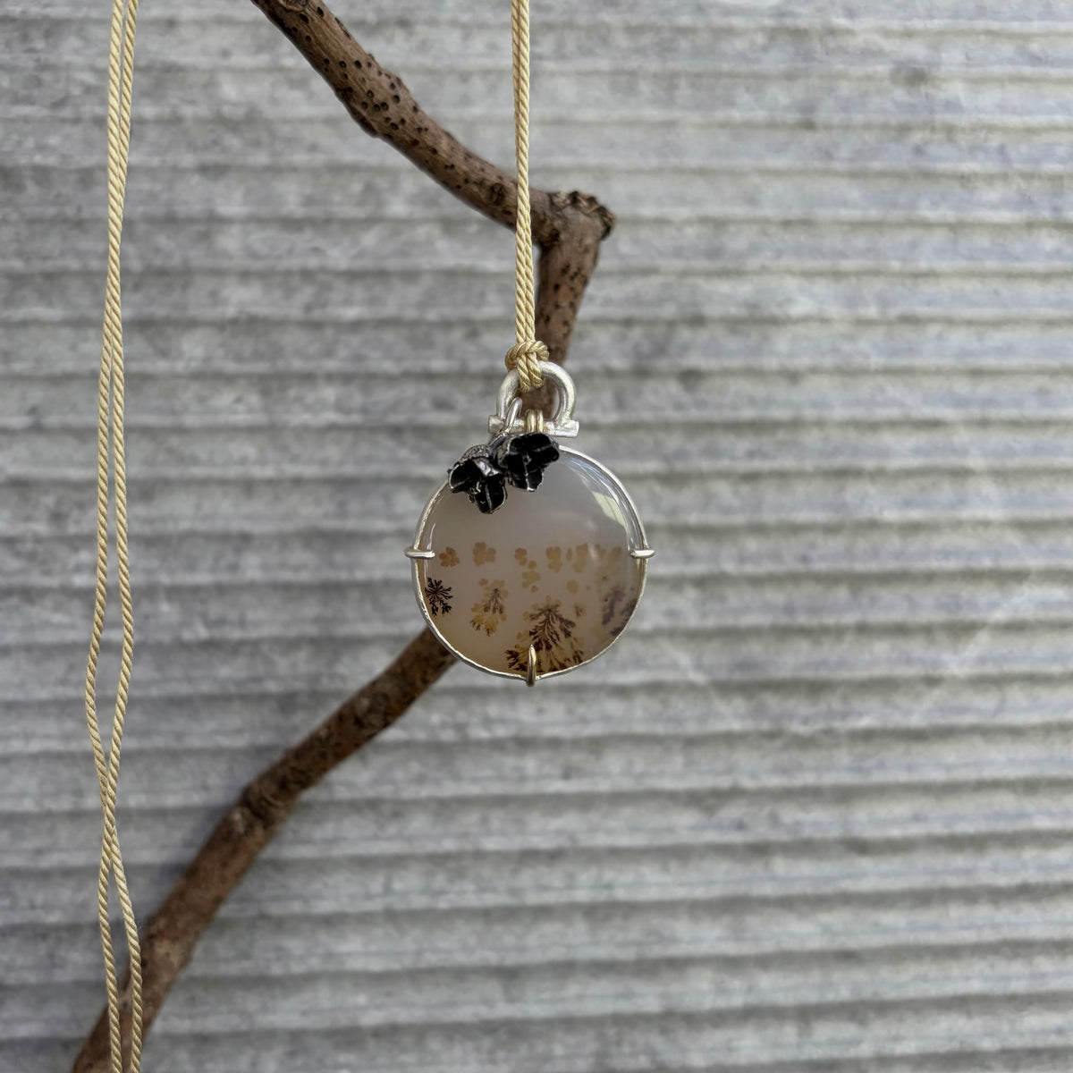 Vertical photograph of a handmade round dendritic agate pendant suspended by a fine golden silk cord,  hanging from a natural  tree branch. The circular gemstone showcases delicate brown branch-like inclusions and is secured in a sterling silver bezel with three structural prongs, including a solid 9ct gold prong at the base. A heavy silver arched bail assembly frames a dark, oxidised cast-from-nature eucalyptus blossom charm, juxtaposed against a neutral grey horizontally ribbed architectural backdrop.