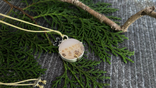 Flat lay photograph of a handmade round dendritic agate pendant resting on green conifer branches nex to a wooden twig. The circular gemstone features natural brown branch-like inclusions and is secured in a thick fine silver bezel with 3 prongs, one of which is a centrally placed 9ct prong. At the top, a structural silver fixed-arch bail is wrapped with a 14ct gold wire coil, suspending a dark, oxidised cast-from-nature eucalyptus blossom cluster charm and a fine golde silk cord.