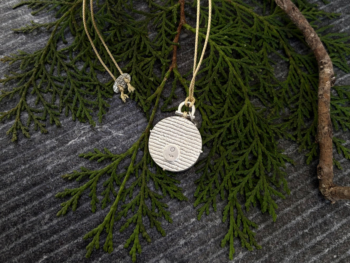 Macro photograph showing the reverse side of a handmade circular sterling silver pendant flat on green pine needles. The thick silver backplate displays a deeply embossed horiontal ribbed texture resembling brutalist industrial architecture. A smooth round disc is positioned off-centre, stamped with a custom maker’s mark and a 925 silver purity hall,ark. At the top, the architectural silver fixed-arch bail assembly attaches cleanly to a golden 3-ply silk cord with a textured oval end disc.