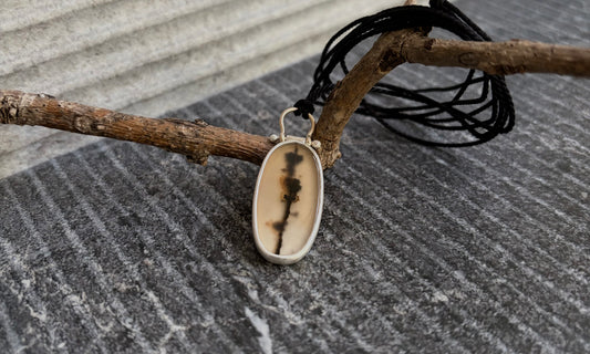 Flat lay photograph of a handmade oval dendritic agate pendant suspended by a black cord, resting on a natural tree branch. The elongated gemstone showcases a single vertical floral stem-like manganese inclusion and is secured in a smooth fine silver bezel setting. At the top, a polished 9ct gold bail forms an arched, articulated hinge loop against a backdrop of grey horizontally ribbed stone .