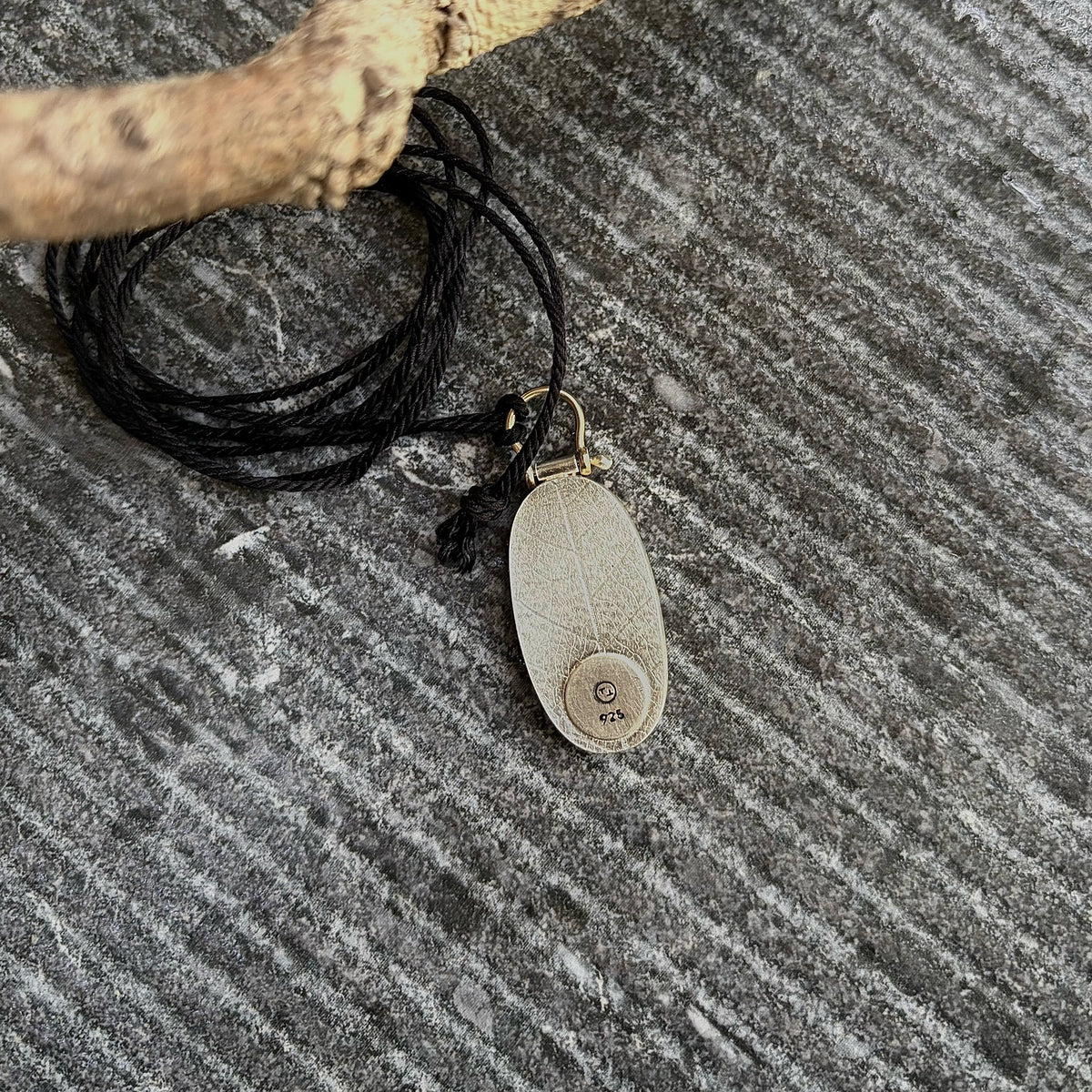 Flat lay photograph showing the reverse side of an oval sterling silver pendant atop a grey textured stone surface. The sterling silver ackplate features an embossed leaf skeleton botanical texture. A smooth, circular disc is positioned off-centre at the base, stamped with a custom studio maker’s mark and 925 hallmark. At the top a golden 9ct gold wire bail loops through a knotted 3-ply black cord.
