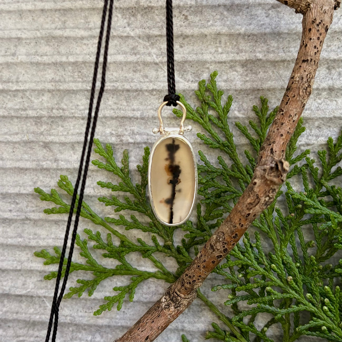 Vertical lifestyle photograph of a handmade oval dendritic agate pendant hanging suspended by a multi-ply black cord from a natural, textured brown tree branch. The elongated translucent gemstone showcases a vertical floral branch-like manganese inclusion at its centre, secured with a smooth fine and sterling silver bezel setting. At the crown, a 9ct gold wire arch bail forms an articulated hinge loop, contrasted against vibrant green conifer foliage and a light grey horizontally ribbed stone background.