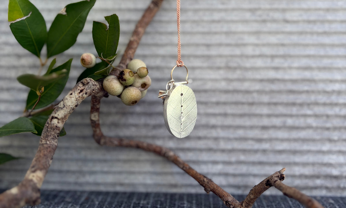 The hand-fabricated sterling silver reverse side of Bespoke Pendant No. 05 showcasing deeply embossed leaf skeleton texture with a line of graduating perforations in the central spine suspended from a branch with foliage.