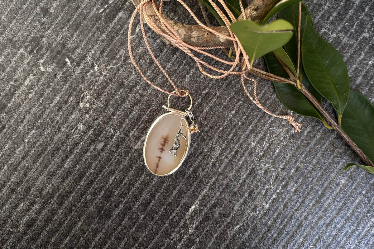Bespoke oval dendritic agate pendant featuring an organic winter branch inclusion, articulated from an arched 9ct gold wire bail with a swinging cast sterling silver gumnut cluster and a cubic zirconia pendant, displayed flat on green leaves.