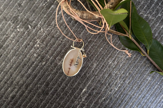 Bespoke oval dendritic agate pendant featuring an organic winter branch inclusion, articulated from an arched 9ct gold wire bail with a swinging cast sterling silver gumnut cluster and a cubic zirconia pendant, displayed flat on green leaves.