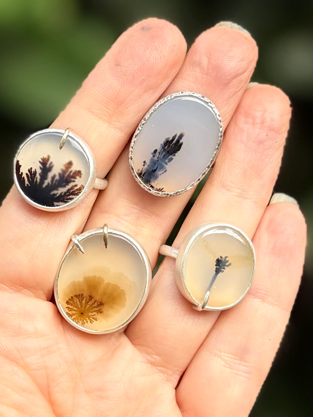 Group view of four bespoke sterling silver dendritic agate rings held in hand, showcasing a collection of translucent dendritic agate stones with unique, organic botanical and moss-like inclusions.
