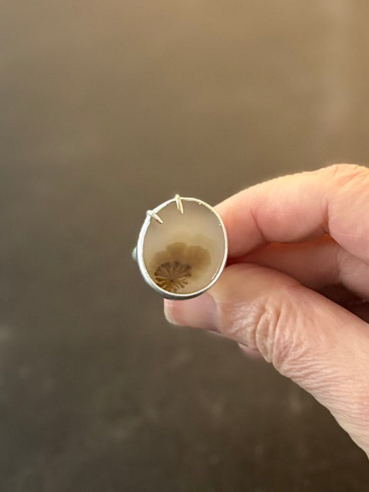 Top view of Bespoke Dendritic Agate Ring held in maker’s hand, showing a translucent agate stone with a graduating dark to light, bloom-like botanical inclusion bezel set with two off-centre claw prongs, one silver and one gold.