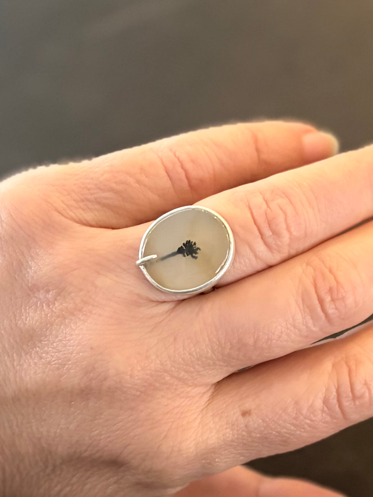 Minimalist dendritic agate ring worn on a finger, displaying the rounded oval dendritic agate stone with its central palm tree inclusion and single off-centre prong against matte stering silver.