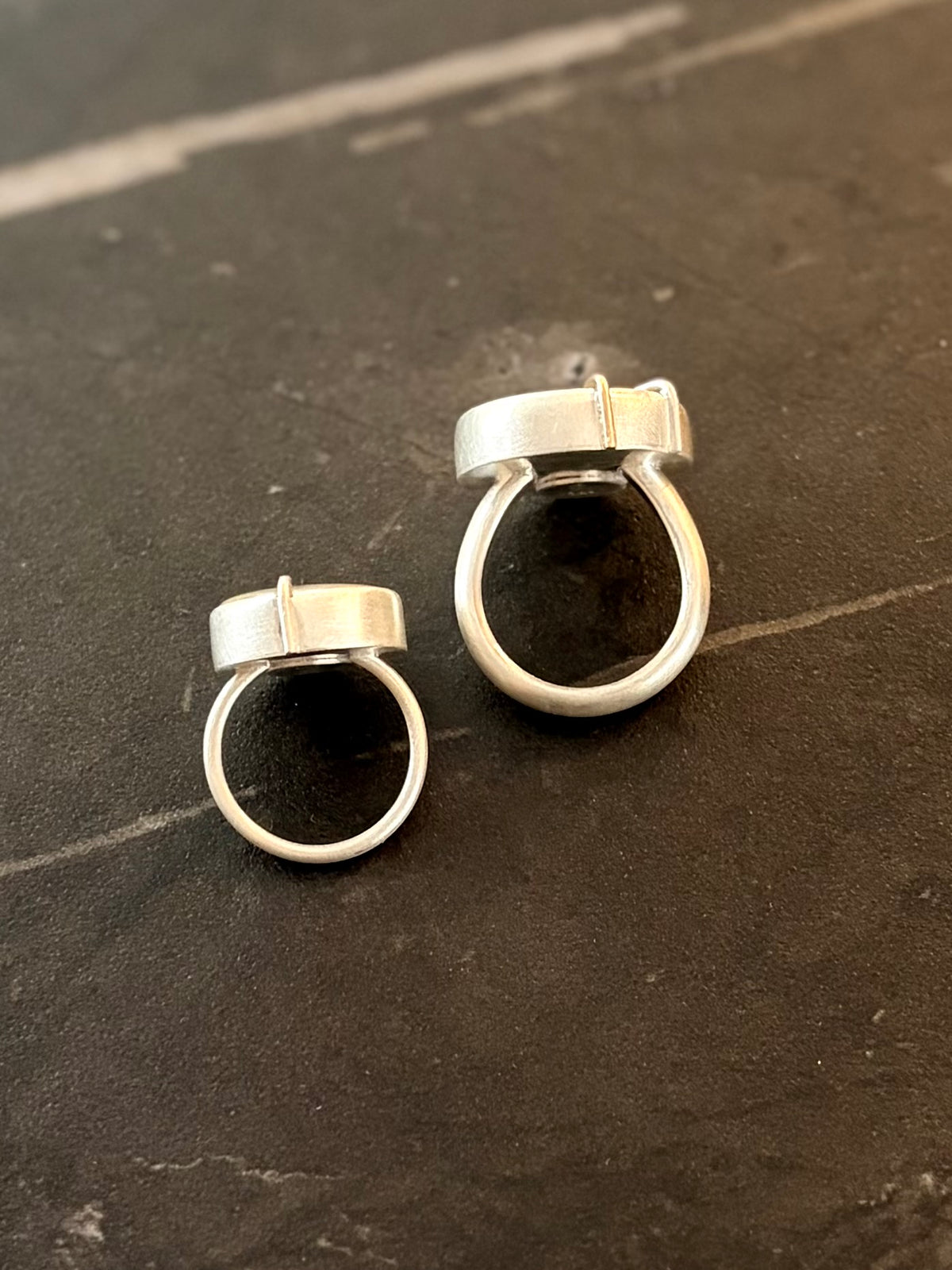 Side profile view of two matte sterling silver rings, highlighting the right ring’s chunky scale! 4.5mm wide band, and specialised anti-spin U-shaped interior profile against a smaller ring with a flat interior band that has a rounder profile.