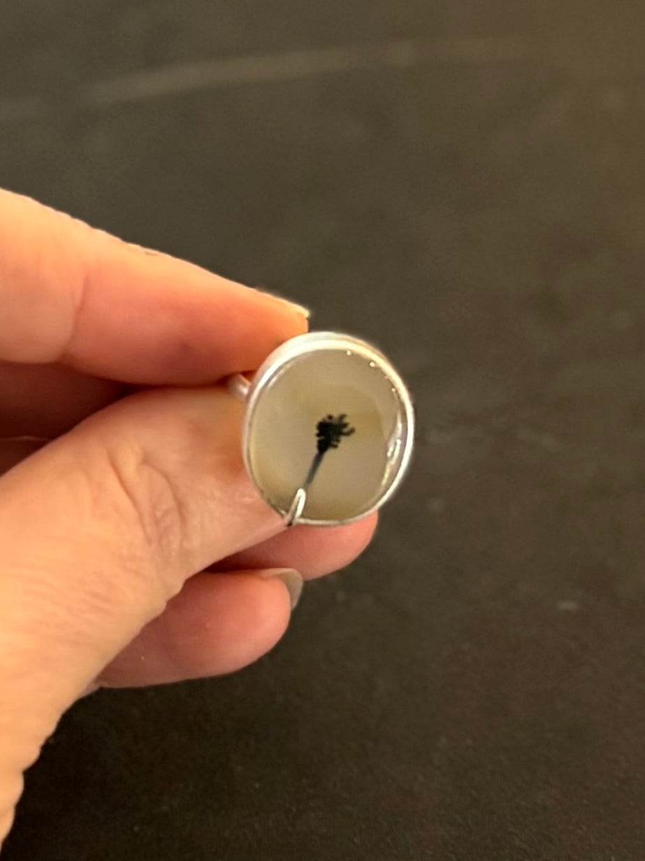 Top view of a bespoke sterling silver dendritic agate ring held in hand, showcasing a rounded oval dedritic agate gemstone with a distint black palm tree silhouette inclusion against a translucent white background.