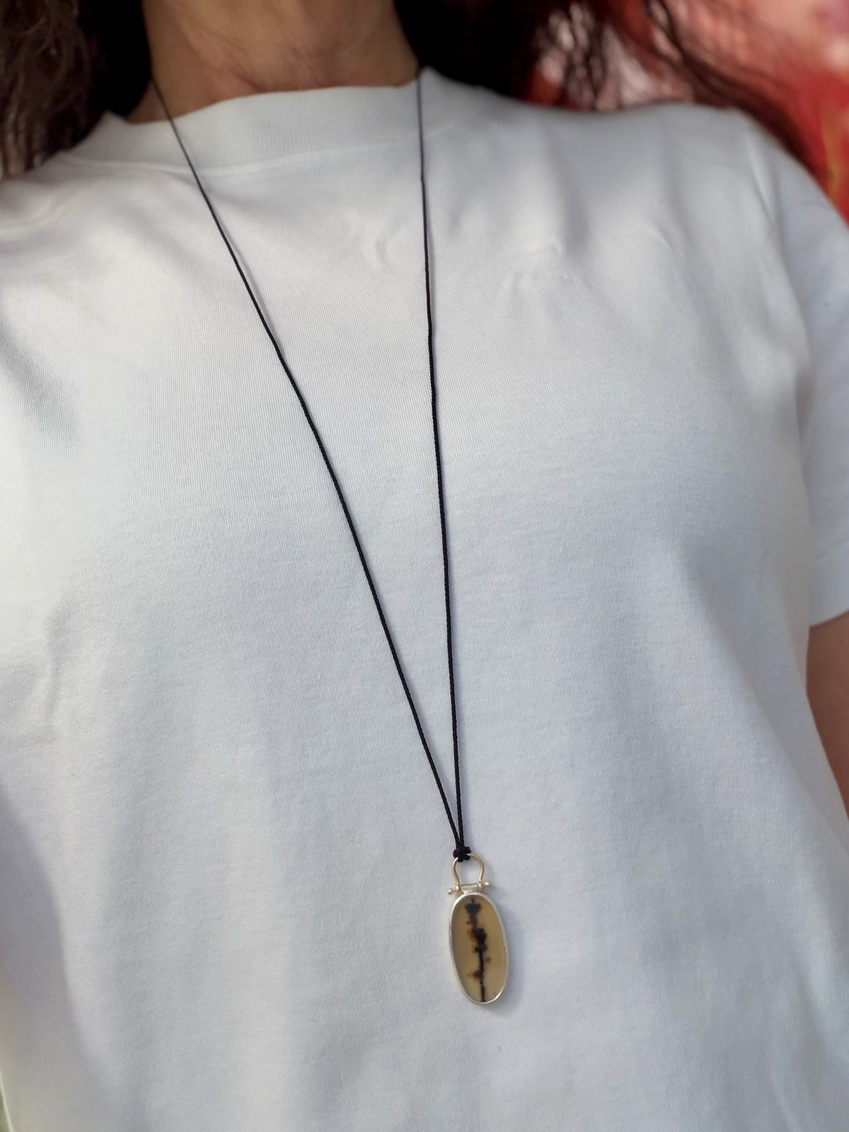 Editorial style shot of the bespoke oval dendritic agate pendant necklace worn over a crisp, structured white crewneck t-shirt. The elongated gemstone, with its prominent vertical branch-like manganese inclusion, is suspended low from a delicate black silk cord contrasting against the heavy white cotton fabric. The smooth fine silver bezel and polished arched 9ct gold bail catch the light, demonstrating the piece’s elegant long drop length, daily wearability, and contemporary aesthetic.