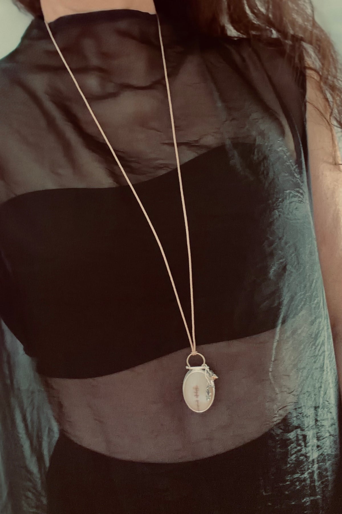 Editorial style shot of the bespoke oval dendritic agate pendant necklace being worn over a sheer, textured black high-neck dress layered over a strapless black bodice. The long, delicate silk cord allows the sculptural pendant to rest elegantly below the chest, showcasing the independent charms - a geometric amber-hued cubic zirconia and a cast gumnut cluster - dangling alongside the silver frame.